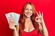 © Krakenimages.com - Young redhead woman holding hong kong dollars banknotes doing ok sign with fingers, smiling friendly gesturing excellent symbol