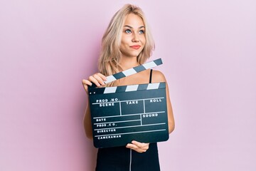 Sticker - Beautiful caucasian blonde girl holding video film clapboard smiling looking to the side and staring away thinking.