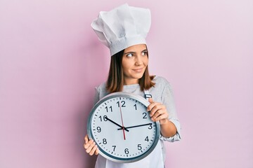 Poster - Young beautiful woman wearing professional cook uniform and hat holding clock smiling looking to the side and staring away thinking.