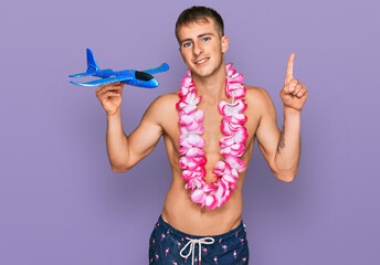 Young blond man wearing swimsuit and hawaiian lei holding airplane toy surprised with an idea or question pointing finger with happy face, number one