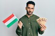 Young hispanic man holding hungary flag and forints relaxed with serious expression on face. simple and natural looking at the camera.