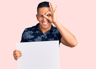 Wall Mural - Young handsome latin man holding cardboard banner with blank space smiling happy doing ok sign with hand on eye looking through fingers