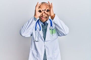 Wall Mural - Hispanic adult man wearing doctor uniform and stethoscope doing ok gesture like binoculars sticking tongue out, eyes looking through fingers. crazy expression.