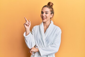 Canvas Print - Young blonde woman wearing bathrobe with a big smile on face, pointing with hand and finger to the side looking at the camera.