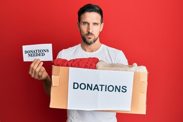 Sticker - Young handsome man volunteer holding box and donations needed paper relaxed with serious expression on face. simple and natural looking at the camera.