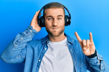 Sticker - Young caucasian man using headphones and doing rock symbol puffing cheeks with funny face. mouth inflated with air, catching air.