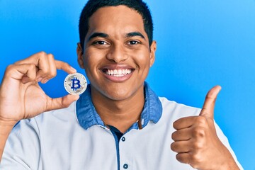 Young handsome hispanic man holding virtual currency bitcoin smiling happy and positive, thumb up doing excellent and approval sign