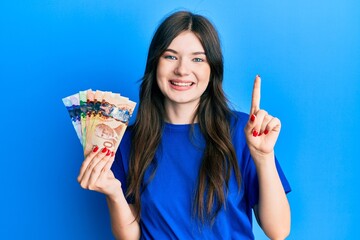 Wall Mural - Young beautiful caucasian girl holding canadian dollars smiling with an idea or question pointing finger with happy face, number one