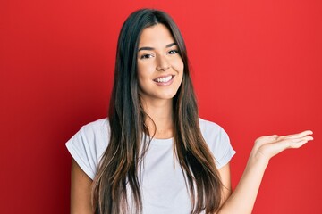 Canvas Print - Young brunette woman wearing casual white tshirt over red background smiling cheerful presenting and pointing with palm of hand looking at the camera.