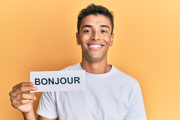 Poster - Young handsome african american man holding bonjour french greeting word looking positive and happy standing and smiling with a confident smile showing teeth