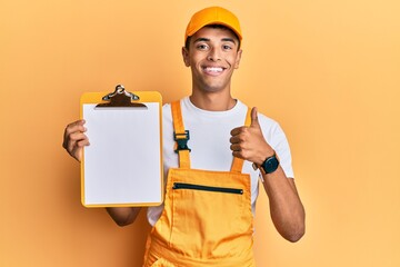 Sticker - Young handsome african american man wearing courier uniform holding clipboard smiling happy and positive, thumb up doing excellent and approval sign