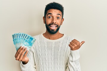 Canvas Print - Handsome hispanic man with beard holding 100 brazilian real banknotes pointing thumb up to the side smiling happy with open mouth