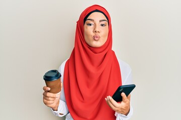 Wall Mural - Young beautiful hispanic girl wearing islamic hijab drinking a coffee usign smartphone looking at the camera blowing a kiss being lovely and sexy. love expression.