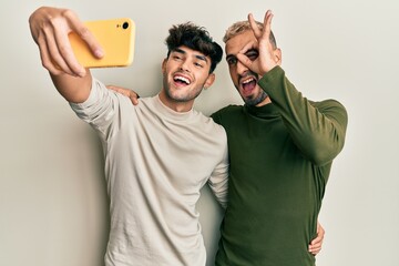 Poster - Homosexual gay couple standing together taking a selfie photo with smartphone smiling happy doing ok sign with hand on eye looking through fingers