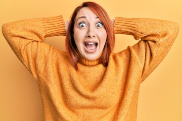 Wall Mural - Beautiful redhead woman wearing casual winter sweater over yellow background crazy and scared with hands on head, afraid and surprised of shock with open mouth