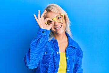 Sticker - Young blonde girl wearing casual clothes and glasses smiling happy doing ok sign with hand on eye looking through fingers