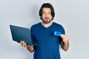 Poster - Middle age caucasian man wearing business style holding laptop and credit card puffing cheeks with funny face. mouth inflated with air, catching air.