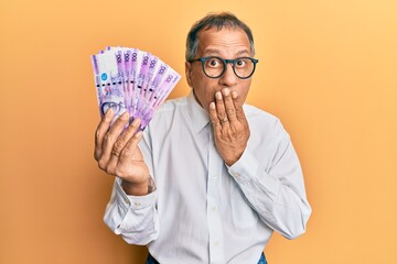 Poster - Middle age indian man holding 100 philippine peso banknotes covering mouth with hand, shocked and afraid for mistake. surprised expression