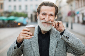 Wall Mural - Mature gentleman in stylish suit smiling on camera, touching grey beard and holding cup of coffee. Urban setting on background. Coffee break outdoors.