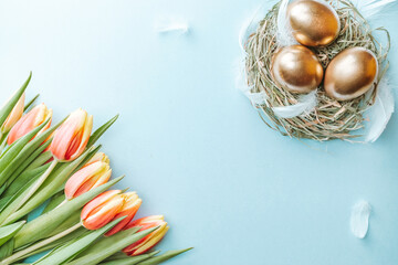 Wall Mural - Easter eggs Golden color in basket with spring tulips, white feathers on pastel blue background in Happy Easter decoration. Festive decoration in flat lay.