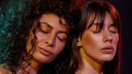 Wall Mural - Beauty portrait of two young brunette female models with professional art makeup posing with eyes closed in neon light isolated over black background