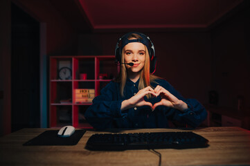 Attractive female gamer in a headset on her head plays video games at home on the computer and streams, looks at the camera with a smile on her face and shows a heart gesture.