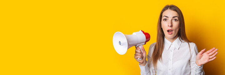 Wall Mural - Surprised young woman in a white shirt holds a megaphone on a yellow background. Hiring concept, ad