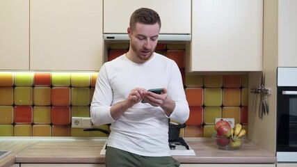 Young guy typing a message on a mobile phone while standing in his kitchen