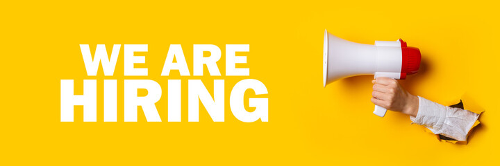 Wall Mural - Female hand sticking out of a hole in the wall holds a megaphone on a yellow background. Added text We are hiring. Hiring concept, help wanted. Banner