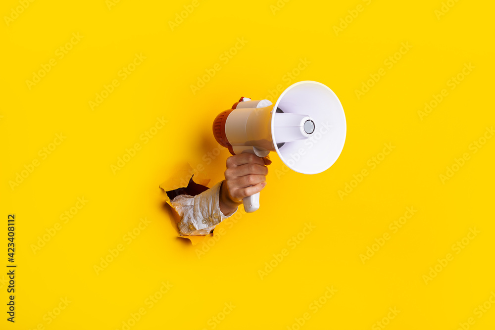 Hand holds a megaphone from a hole in the wall on a yellow background. Concept of hiring, advertising something. Banner - obrazy, fototapety, plakaty 