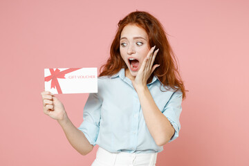 Young shocked surprised amazed impressed happy ginger student redhead woman 20s in blue shirt holding gift voucher flyer mock up holding face isolated on pastel pink color background studio portrait