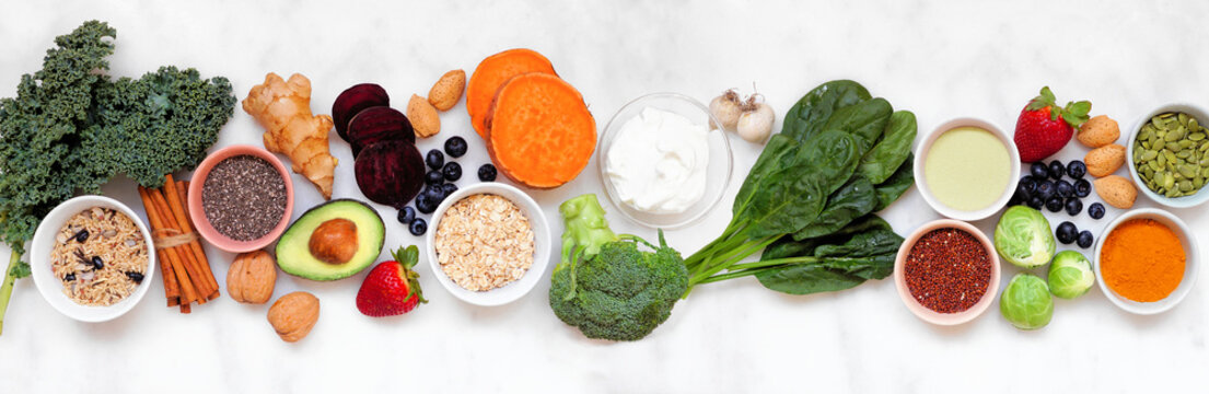 Set of healthy food ingredients. Top view table scene on a white marble banner background. Super food concept with green vegetables, berries, whole grains, seeds, spices and nutritious items.