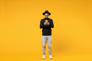 Wall Mural - Full length young friendly smiling happy american african man 20s in stylish black shirt hat eyeglasses using mobile cell phone chat in social network isolated on yellow background studio portrait