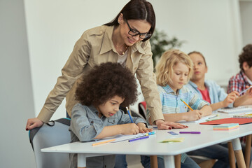 Caring young female teacher in glasses helping little schoolboy. Kids sitting at the table, studying in elementary school classroom