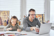 © boytsov - Busy family at home during coronavirus. Child girl drawing next to dad who is working in home office with laptop and headphones. Babysitting during covid-19 lockdown. Front view.