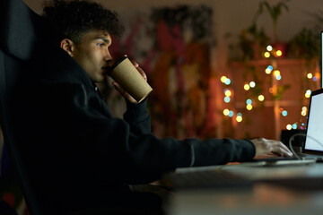 Wall Mural - Coffee break. Side view of young guy with piercing drinking coffee, sitting at the table in front of many computer monitors and working from home late at night