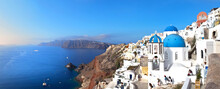 Greece. Santorini Island. Panoramic View On Beautiful Oia Village With White Cycladic Houses And Traditional Churches With Blue Domes At Sunny Day Against Backdrop Of Sea. Summer Holidays And Travel