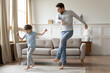 Happy young Caucasian father and small preschooler son dance together in living room on family leisure weekend. Overjoyed dad and little boy child have fun jump and move, celebrate relocation.