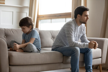 Wall Mural - Angry young Caucasian father and little son sit separately on couch at home have family misunderstanding or fight. Unhappy mad dad and small boy child ignore avoid talk. Generation gap concept.