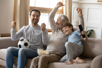 Wall Mural - Smiling three generations of Caucasian men have fun cheering watching football on TV with popcorn snacks. Overjoyed boy child enjoy television with young father and mature grandfather. Fan concept.