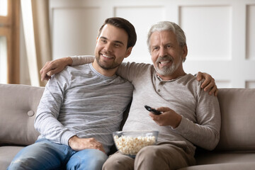 Wall Mural - Smiling senior 60s Caucasian father and adult son sit relax on sofa at home watching TV on weekend together. Happy older dad and grownup millennial male child rest enjoy program on television.