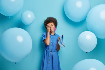 Wall Mural - Pleased dark skinned curly woman makes face palm smiles broadly holds high heel shoes wears fashionable dress poses against blue background around inflated balloons. Festive occasion concept