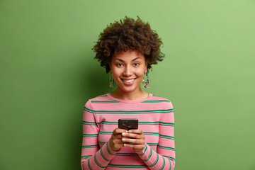 Beautiful young African American woman smiles positively sends text messages via smartphone enjoys online communication being always in touch wears striped jumper isolated over green background