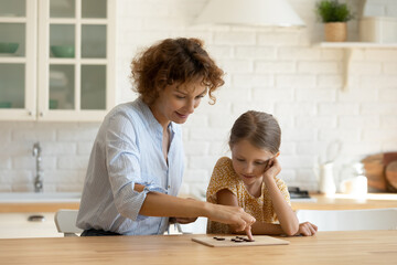 Wall Mural - Caring young Caucasian mother and little daughter have fun engaged in interesting wooden board game. Happy mom and small 9s girl child feel playful enjoy chess develop logic or strategic thinking.