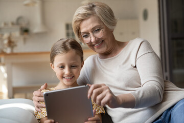 Wall Mural - Smiling mature grandmother and small granddaughter look at tablet screen talk speak on video call online. Happy senior granny and little girl child have fun use modern pad device at home on weekend.