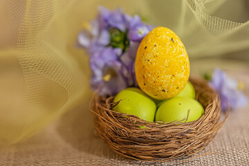 Wall Mural - Large yellow and small green eggs in the nest and a transparent yellow cloth on the burlap. Blurred airy background with flowers.