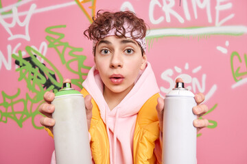 Poster - Photo of surprised curly haired handsome hipster guy uses aerosol spray for drawing graffiti marks public place looks wondered at camera poses against colorful painted wall. Visual communication