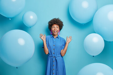 Wall Mural - Indoor shot of positive curly haired Afro American woman raises palms dressed in festive dress looks above isolated over blue background with inflated balloons around enjoys birthday celebration