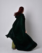 full length portrait of red haired girl wearing celtic, green medieval gown with fantasy velvet cloak. Standing pose isolated against a studio background.