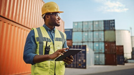 Smiling Portrait of a Handsome African American Black Industrial Engineer in Yellow Hard Hat and Safety Vest Working on Tablet Computer. Foreman or Supervisor in Container Terminal.